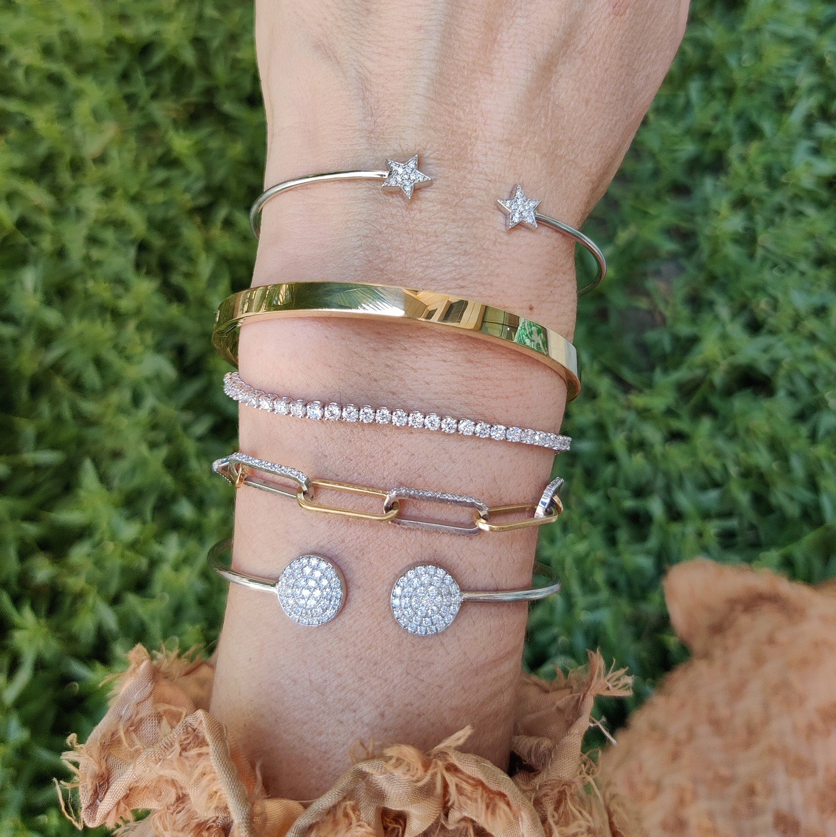 Hand adorned with various bracelets, including a sparkling diamond tennis bracelet, set against a green background.