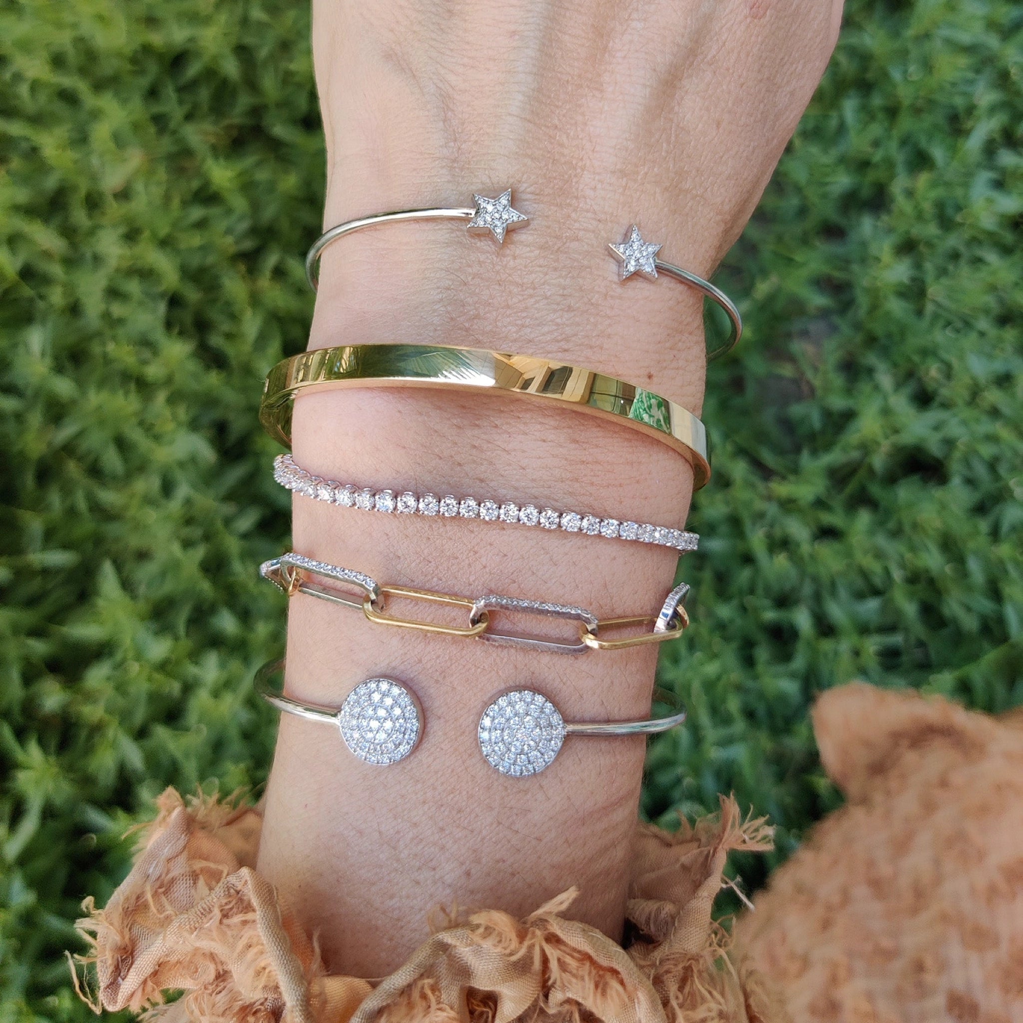 Hand adorned with various bracelets, including a sparkling diamond tennis bracelet, set against a green background.