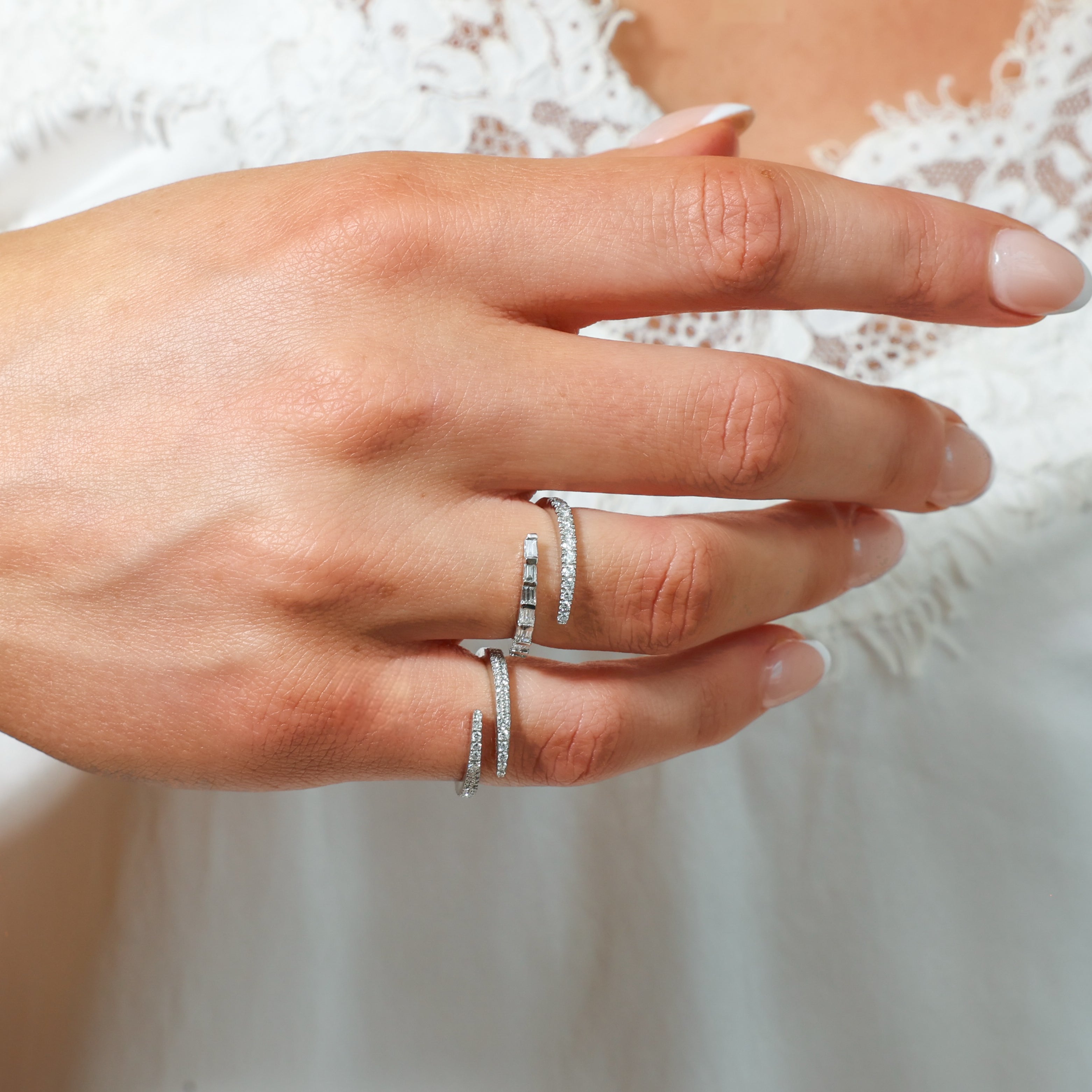 A close-up of a hand wearing diamond wrap rings, styled with a white lace garment in the background.