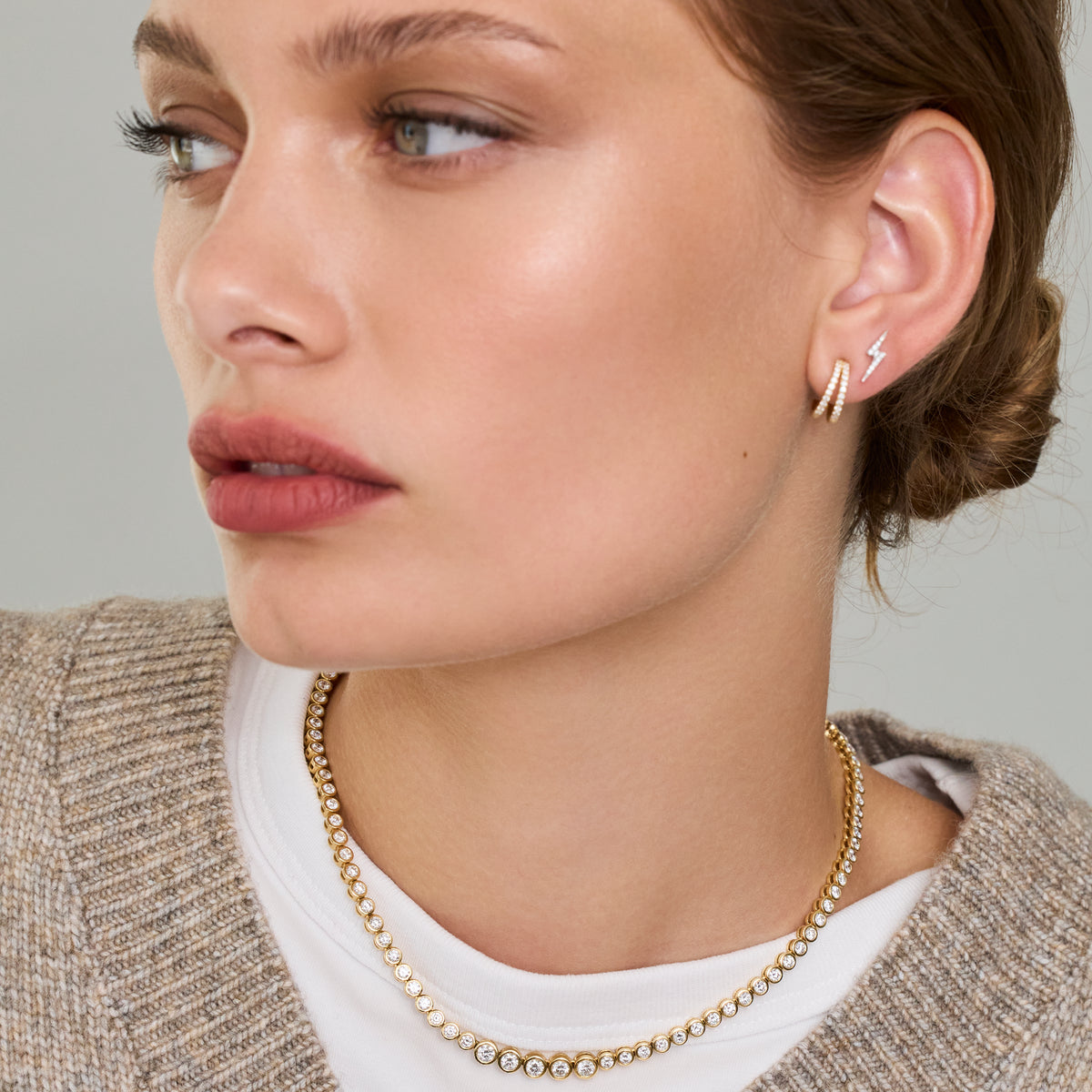 Close-up of a woman wearing a delicate small gold diamond hoop earring with a white stone, against a soft gray background.