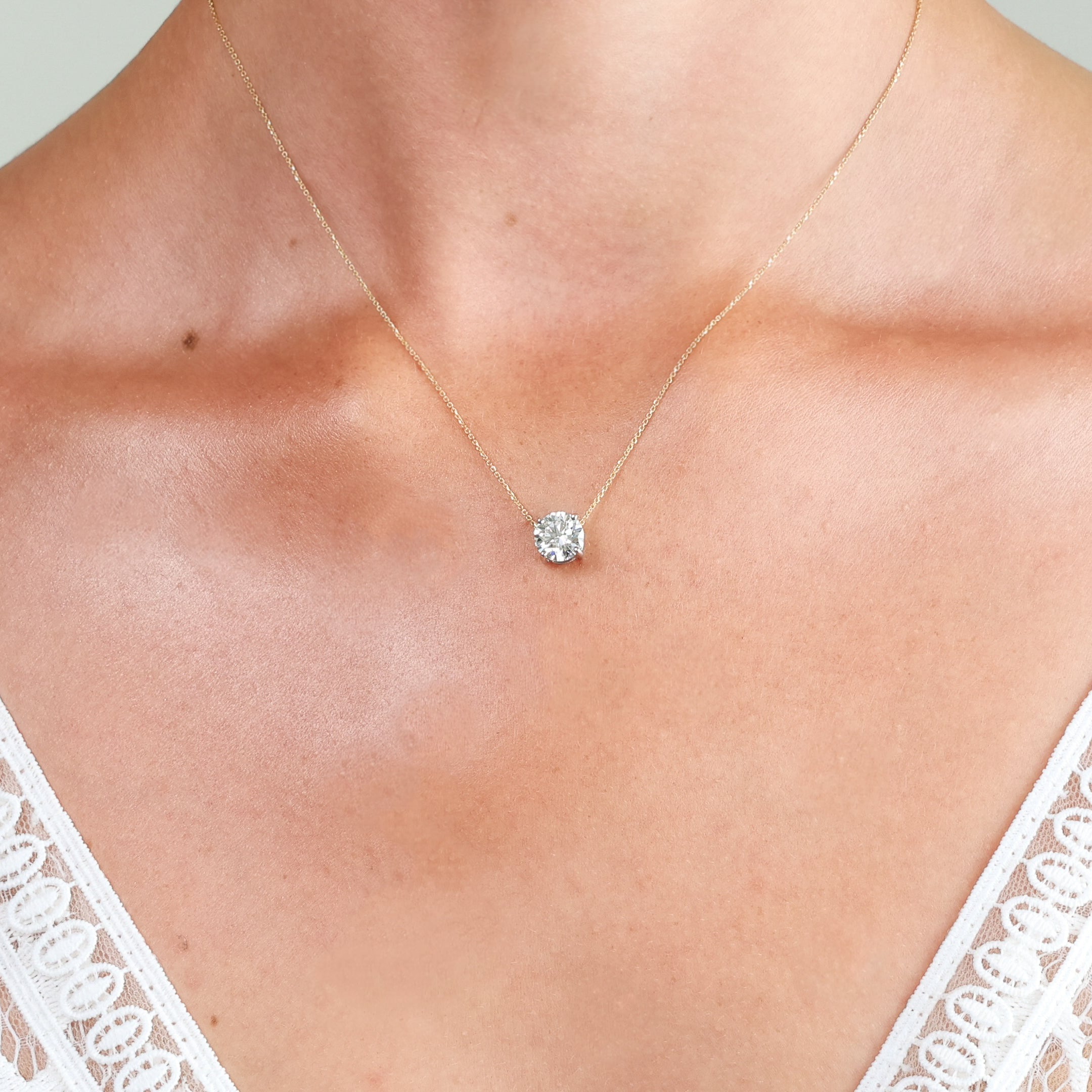 A delicate gold chain necklace with a single round-cut diamond pendant, shown worn on a model's neck above a white lace top.