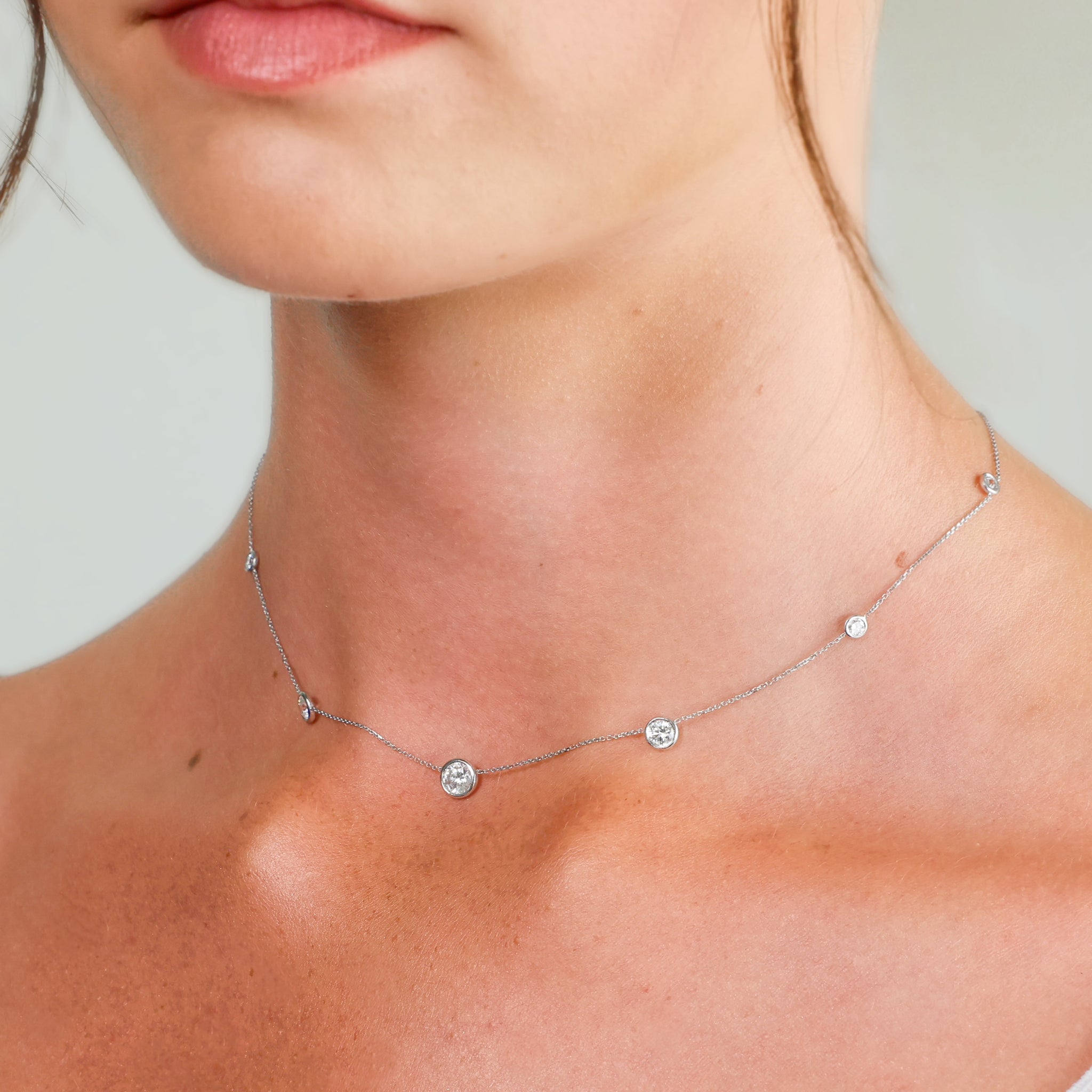 A close-up of a delicate diamond necklace with five small round bezel set diamonds worn on a model's neck.
