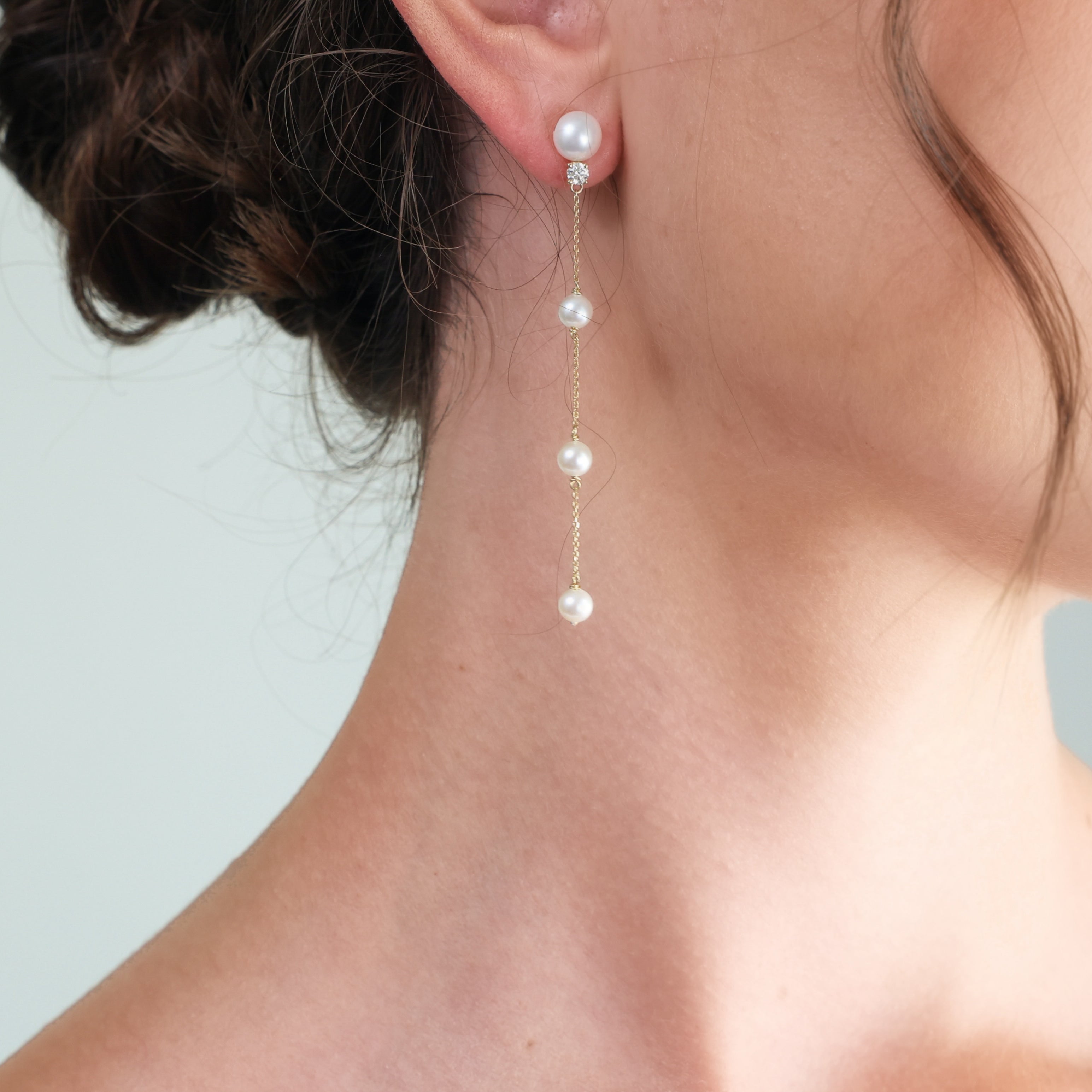 A close-up of a model's ear and neck wearing a delicate pearl drop earring. The earring features a gold chain with four white pearl beads hanging vertically.