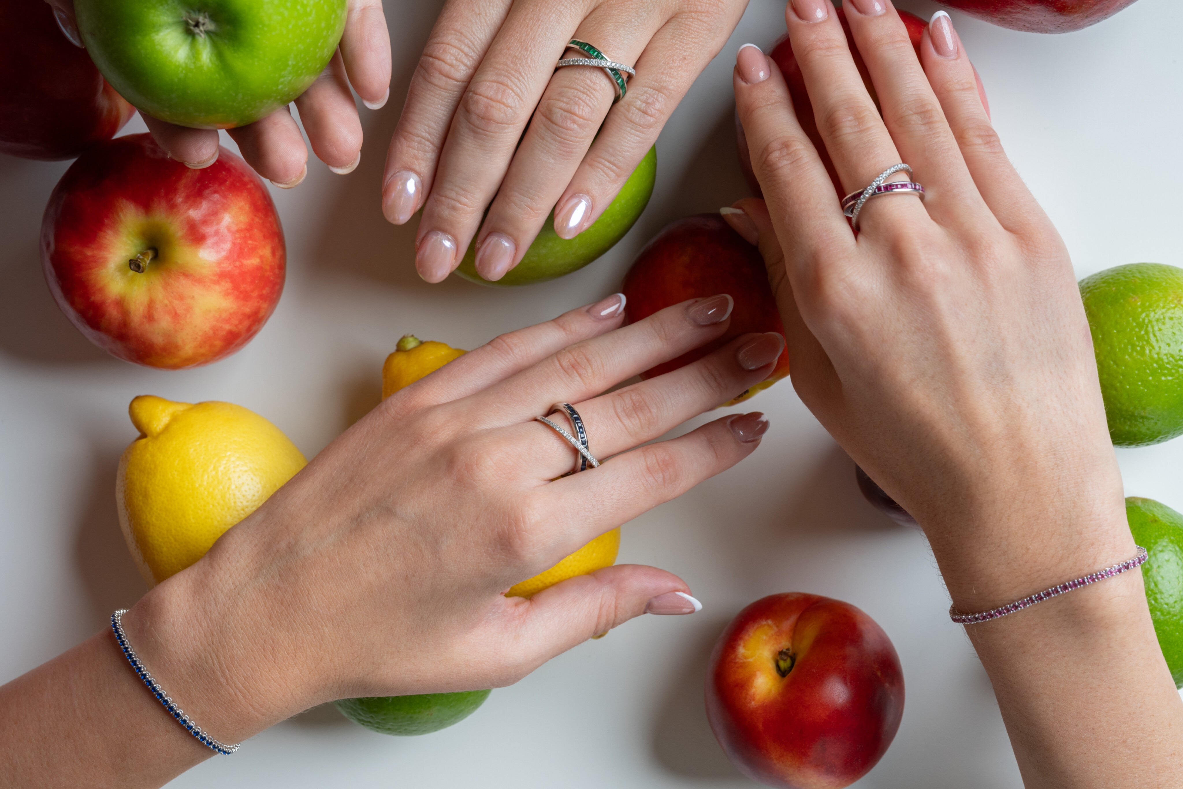 Two hands adorned with gemstone tennis bracelets and rings surrounded by colourful fruits on a white surface.