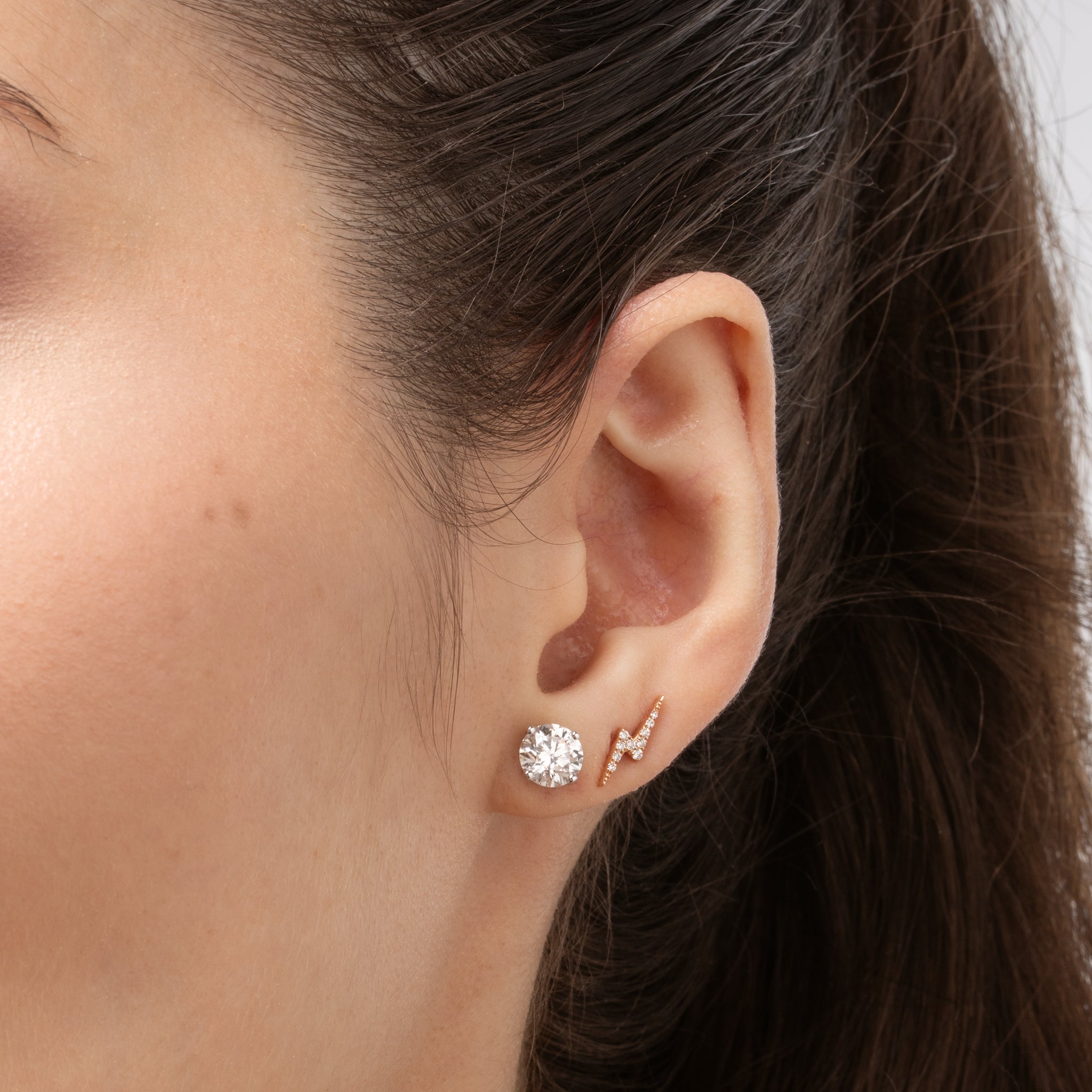 Close-up of a woman's ear showcasing lightning bolt earrings set with certified diamonds, complemented by a sparkling round diamond stud earring.