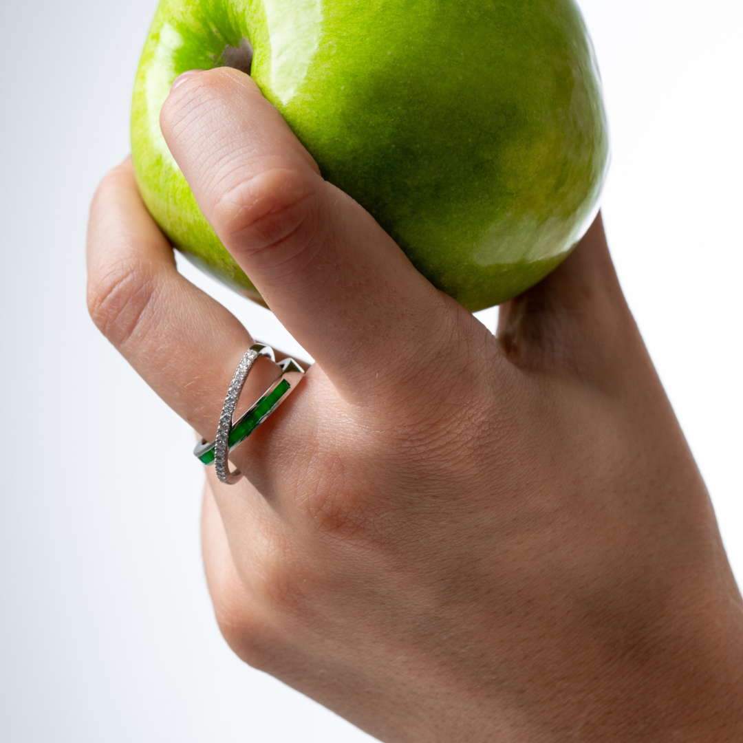 Emerald Criss Cross Ring featuring green emeralds and diamonds worn on a hand holding a green apple.