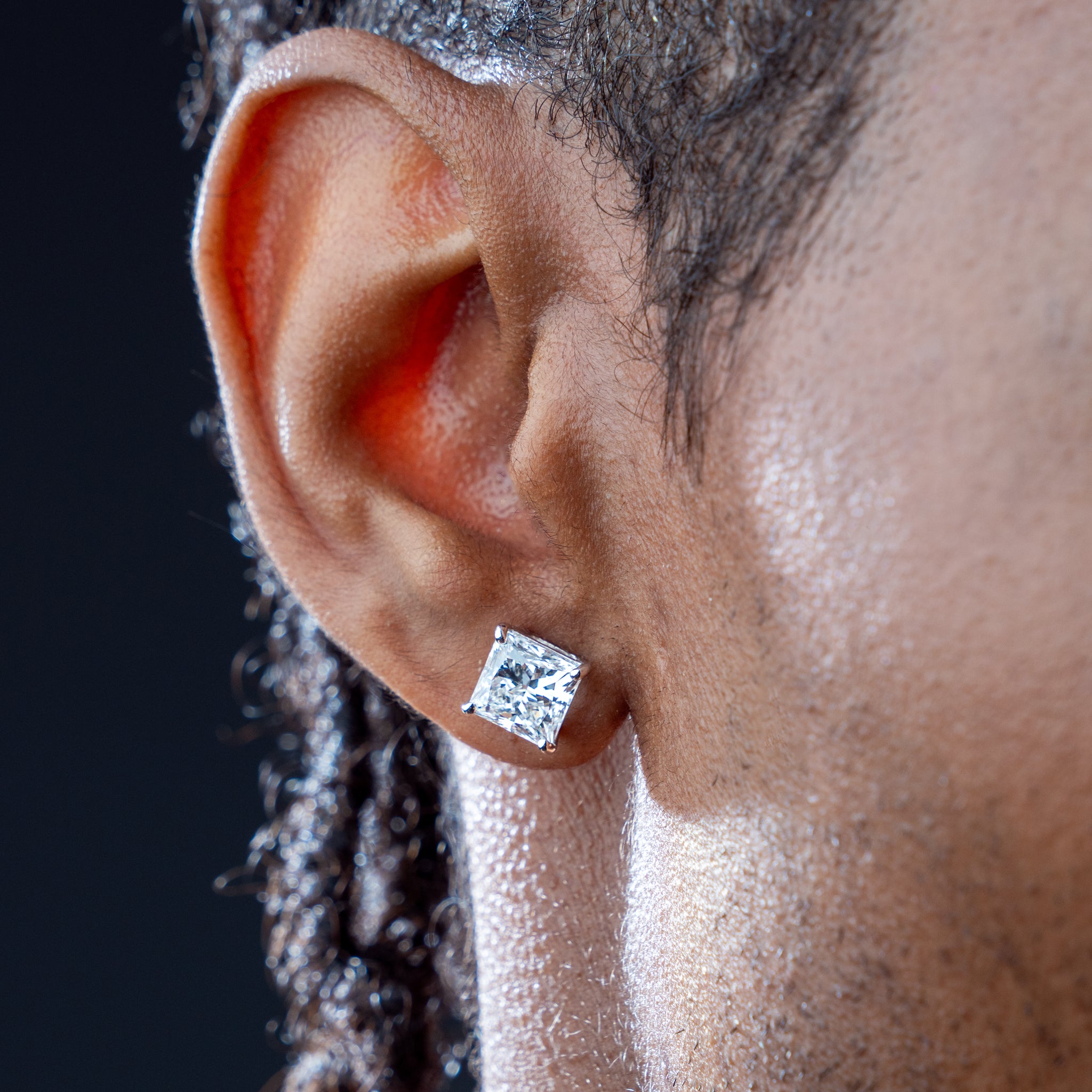 Close-up of an ear wearing a princess cut diamond earring with a dark background