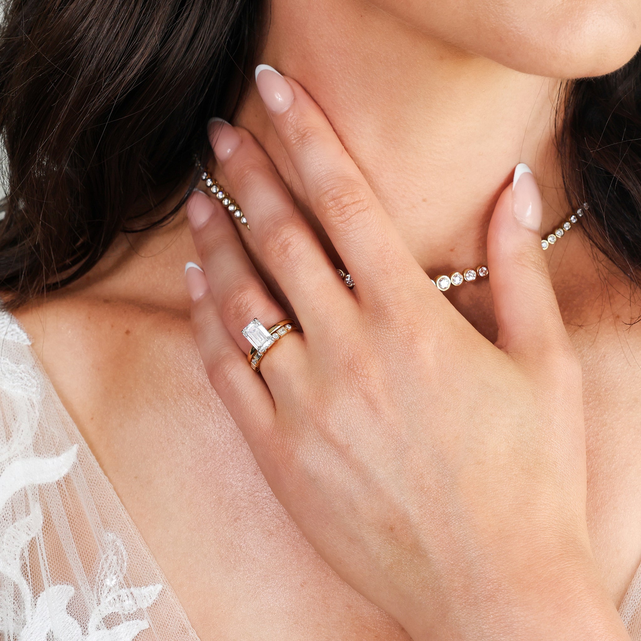 A close-up lifestyle shot of a woman wearing a diamond engagement ring with a round brilliant cut and an emerald cut eternity ring with matching delicate gold and diamond necklaces