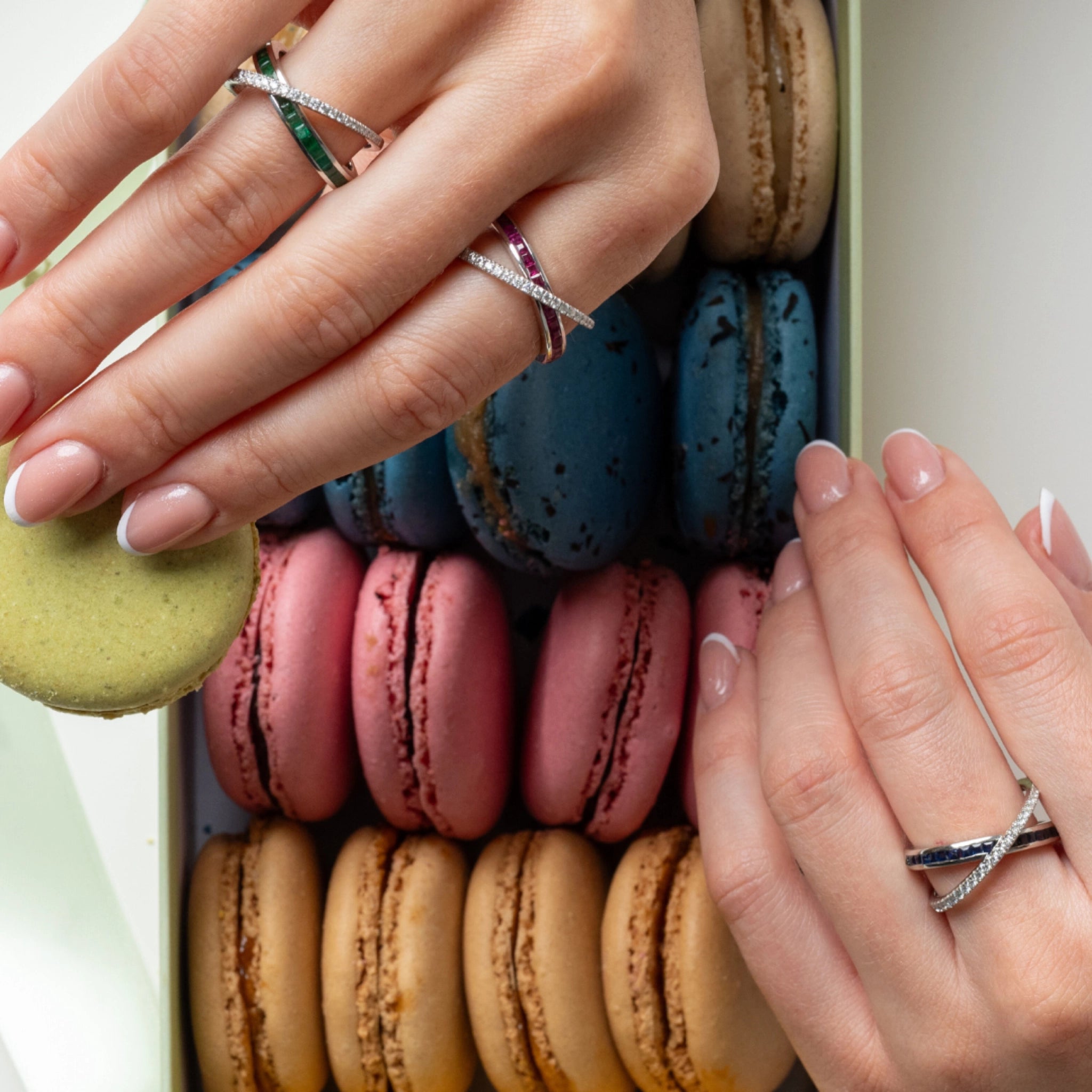 Two hands adorned with a sapphire criss cross ring, elegantly placed over a box of colourful macarons.