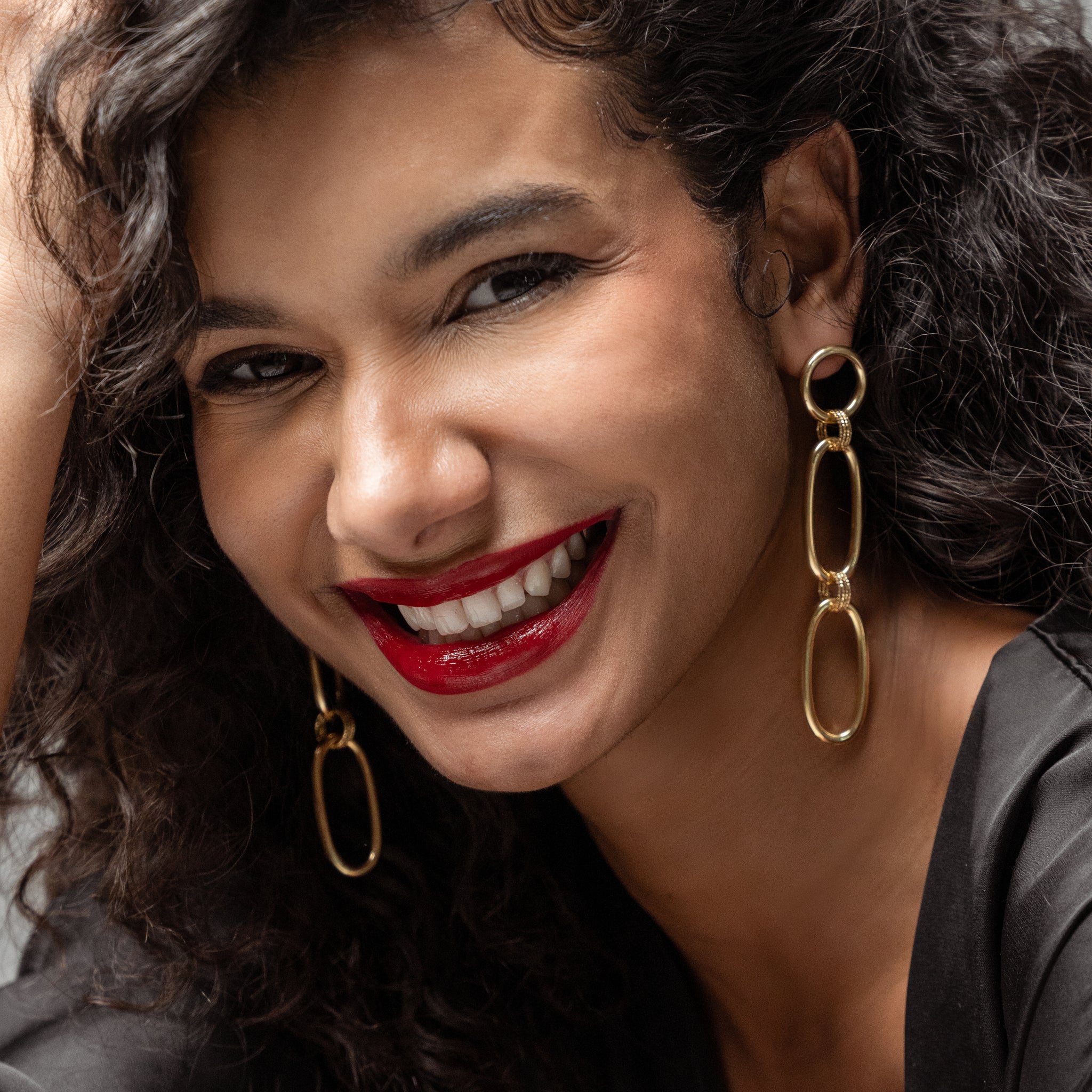 Woman with curly hair wearing gold chain earrings and red lipstick, smiling.