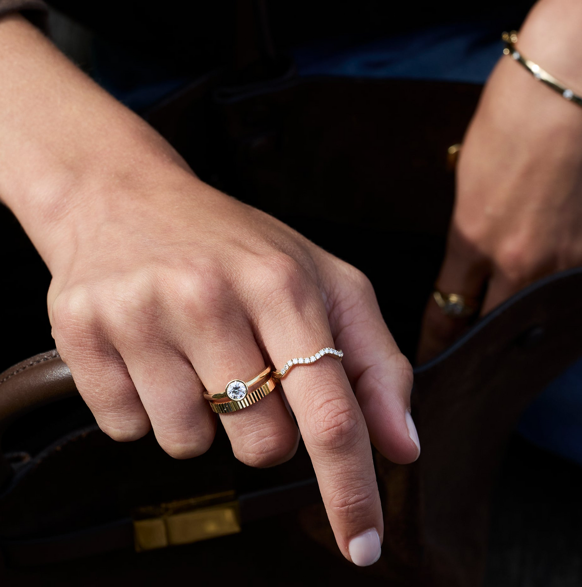 A close-up lifestyle shot of a person's hand wearing three delicate rings: a wave diamond ring with a round-cut bezel set round diamond ring and a gold fluted ring.