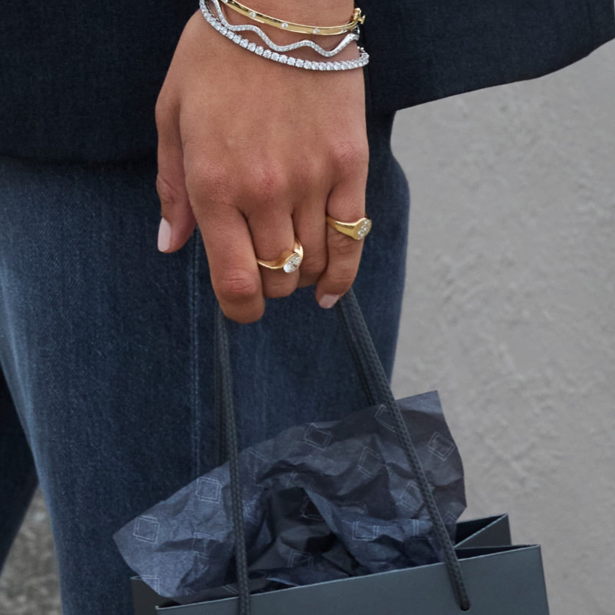 A close-up of a person's hand holding a black shopping bag wearing multiple gold and diamond tesnnis bracelets and diamond rings, dressed in dark blue jeans and a dark top against a light gray wall.
