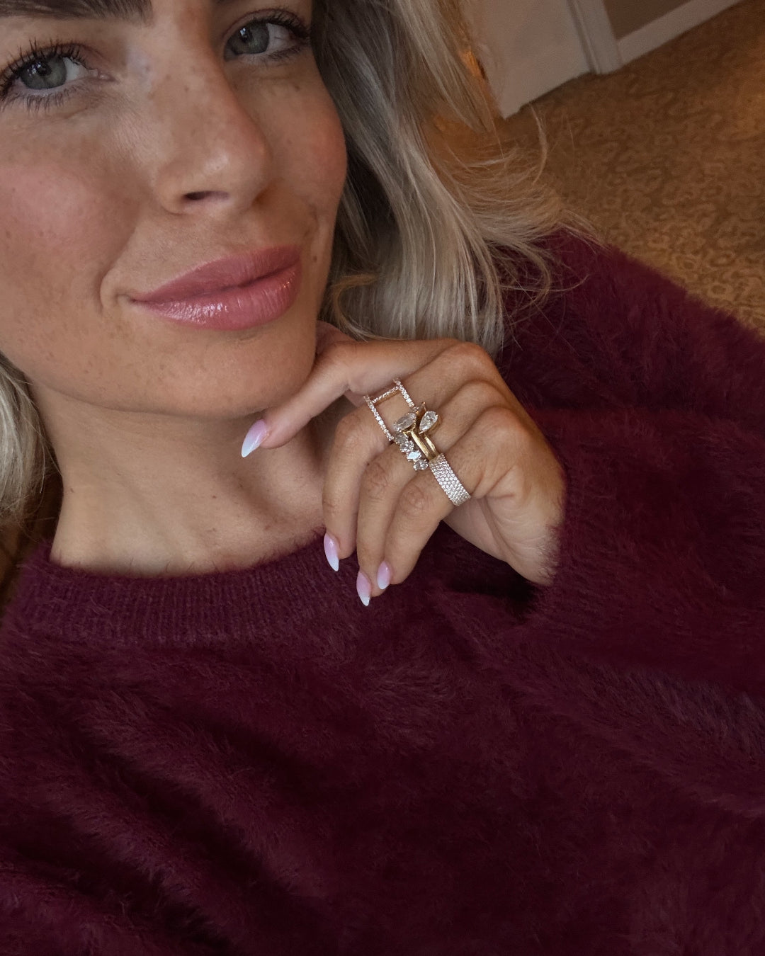 A woman showcases her stylish rings, including a four row pavé diamond ring, wearing a plush maroon sweater.