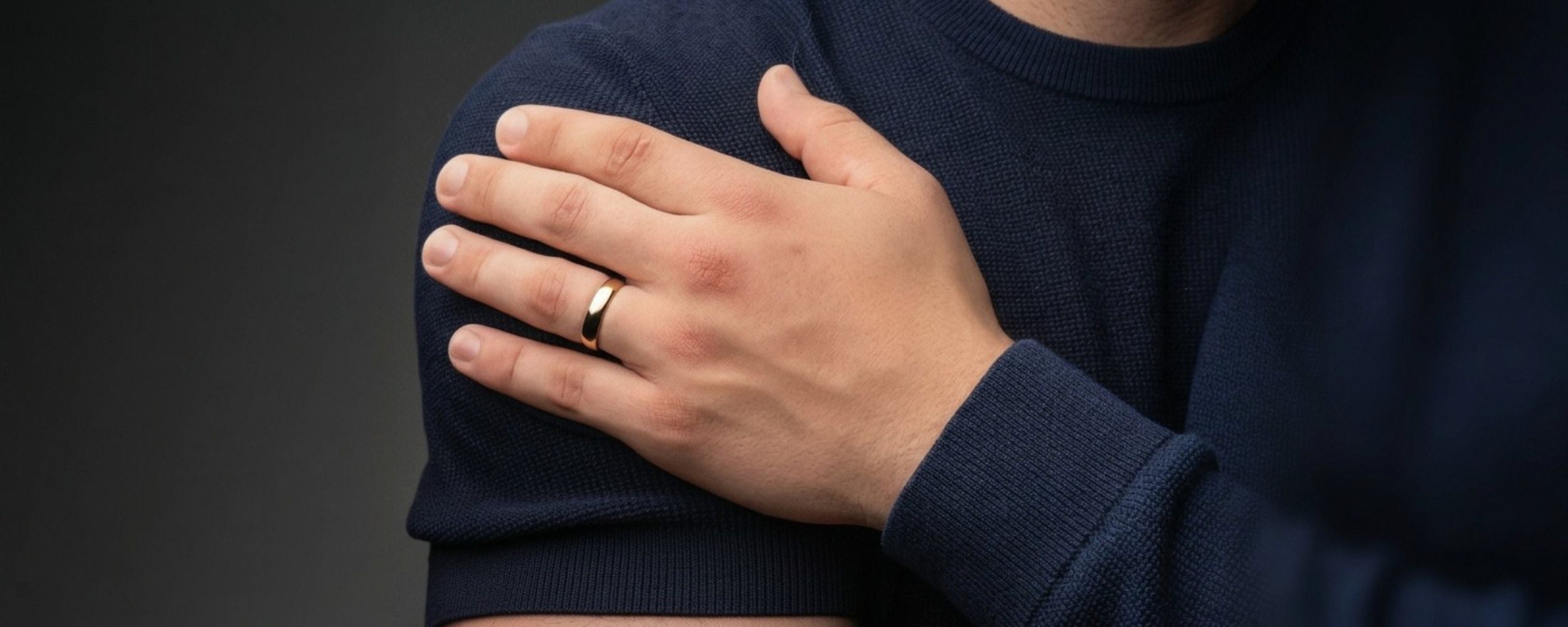 A close-up of a man wearing a gold wedding ring on his ring finger, with his hand resting on his shoulder.