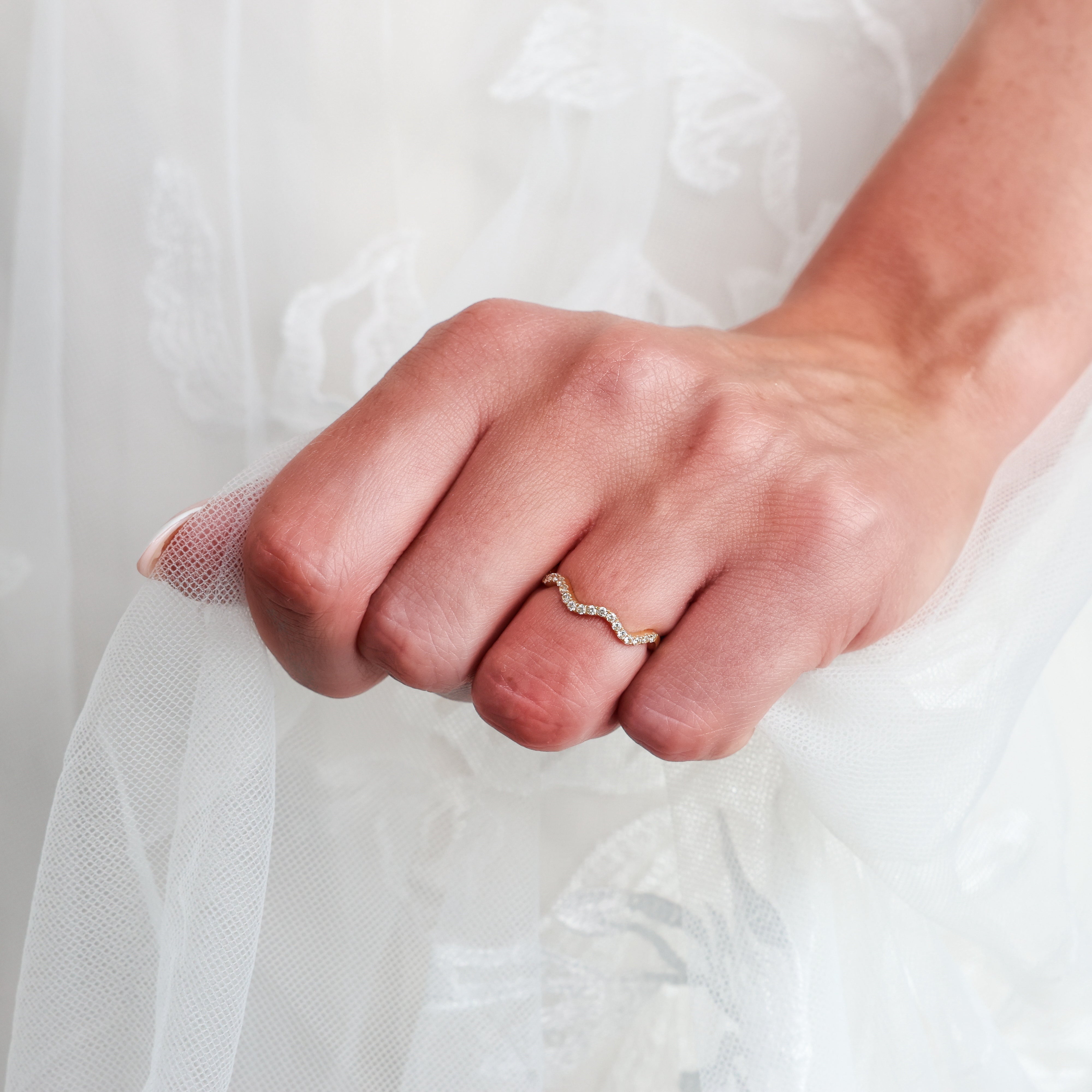 A close-up of a person's hand wearing a delicate wavy gold diamond ring set against a soft white lace and tulle wedding dress
