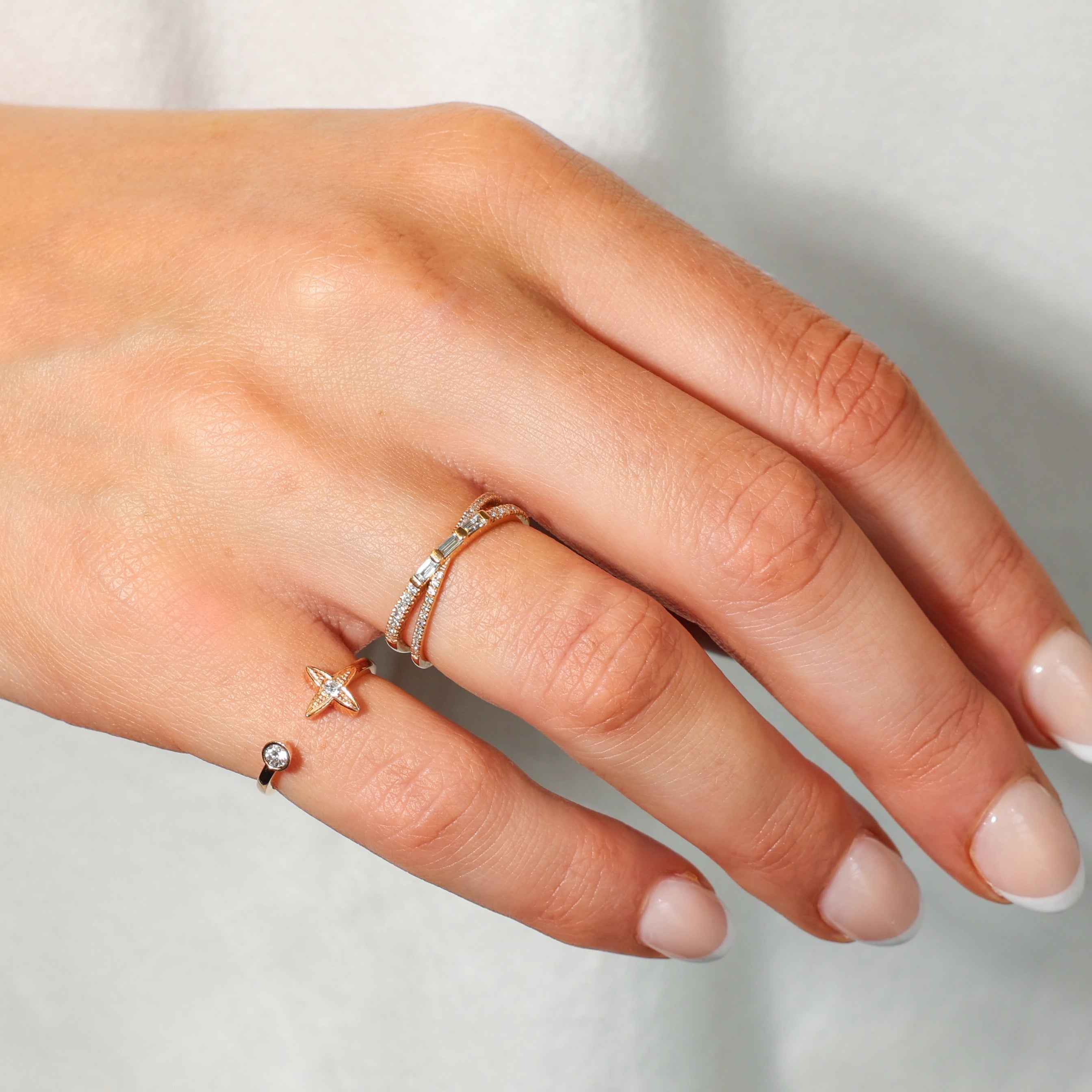 A close-up of a hand wearing delicate diamond rings: a small diamond criss cross ring, a rose gold ring with a small diamond star  