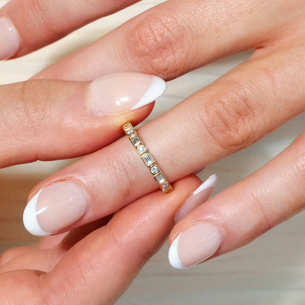 A close-up of a hand wearing a delicate gold eternity ring set with diamonds, featuring a classic French manicure.