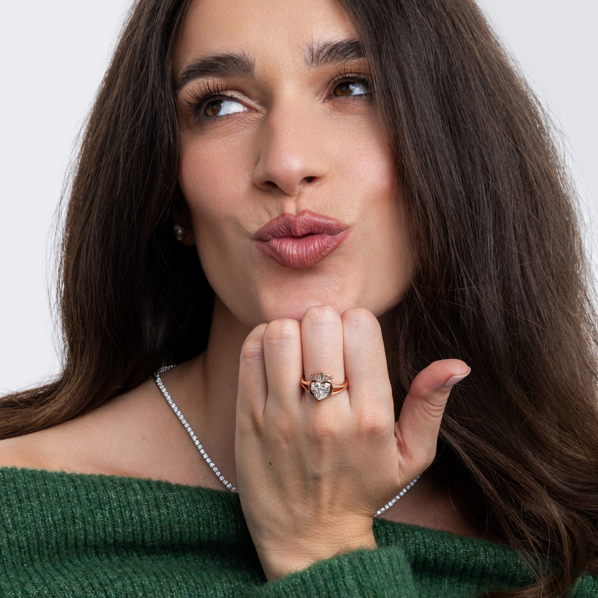 Woman showcasing a Claddagh ring with a thoughtful expression, wearing a green sweater and sparkling necklace.