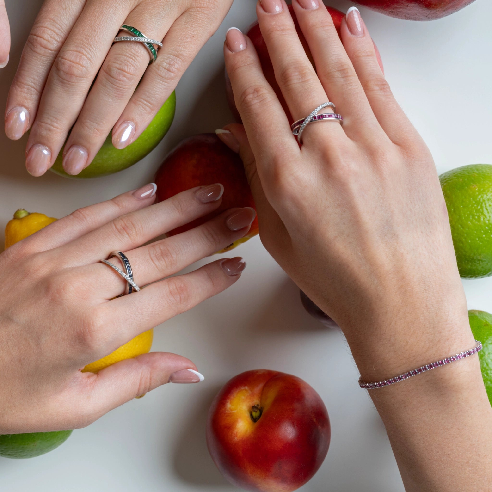 Two hands adorned with gemstone tennis bracelets and rings surrounded by colourful fruits on a white surface.