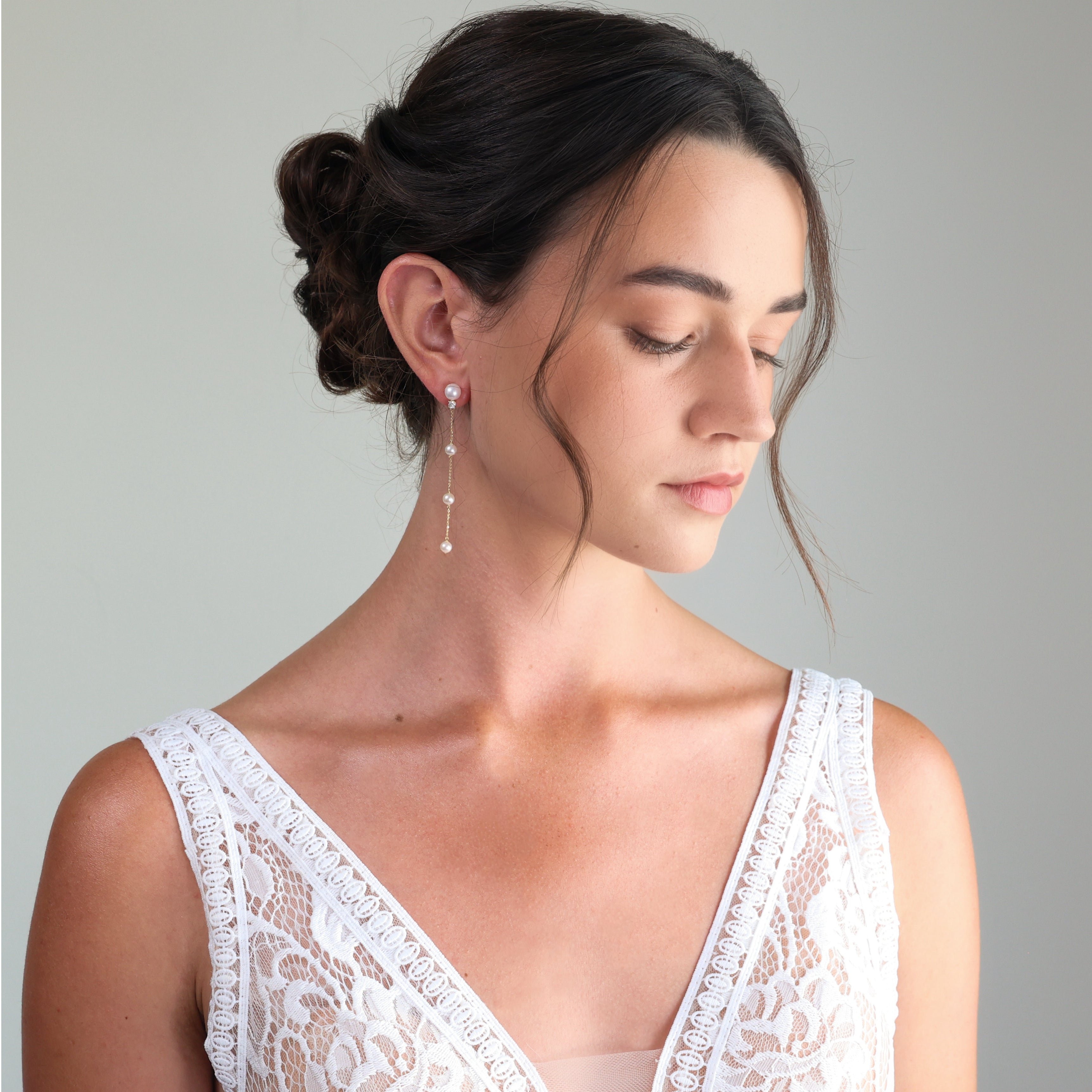 A woman with her hair in a low bun wears a delicate drop earring with white pearls. She is wearing a white lace top against a neutral gray background.