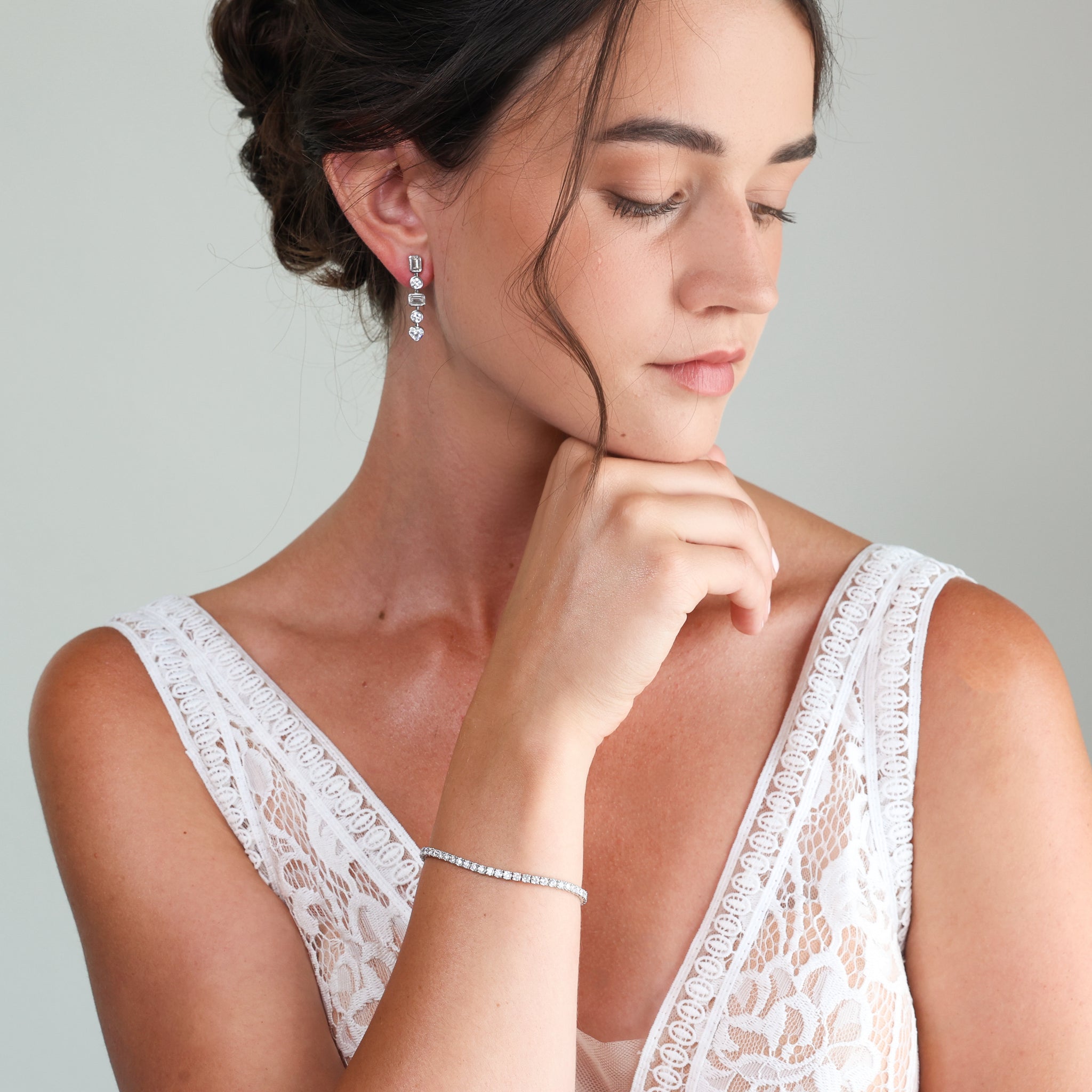 A woman wearing a diamond tennis bracelet on her wrist and a matching diamond drop earring, dressed in a white wedding dress against a soft gray background.