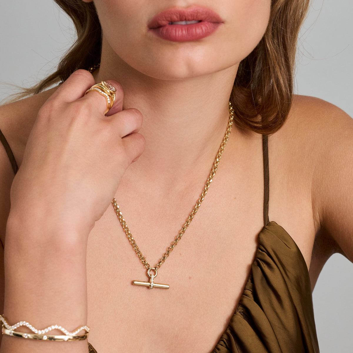 A close-up of a woman wearing a delicate gold T-bar necklace with a small horizontal bar pendant, along with a gold ring and a wave diamond bracelet, against a neutral gray background.
