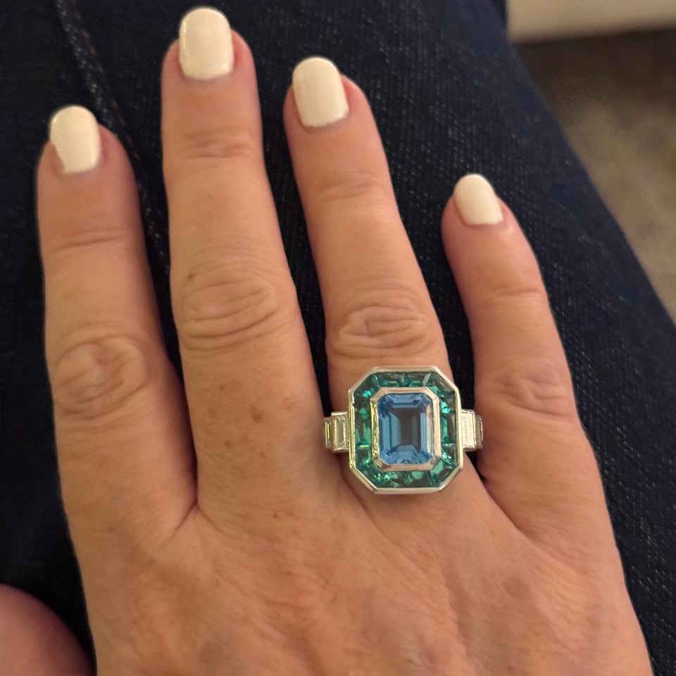An Art Deco style ring featuring a large light blue topaz gemstone surrounded by a double halo of emerald cut green emeralds 