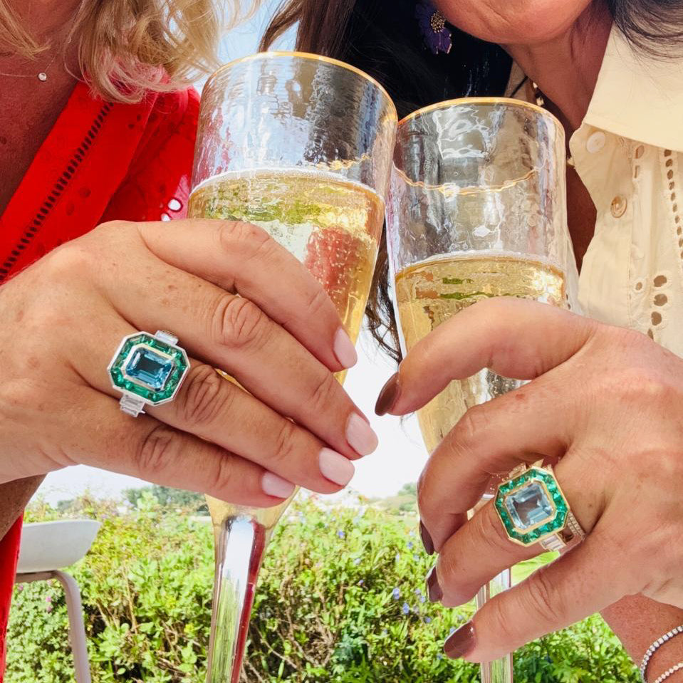 An Art Deco style ring featuring a large light blue topaz gemstone surrounded by a double halo of emerald cut green emeralds, set in a polished yellow gold band with baguette cut side diamonds
