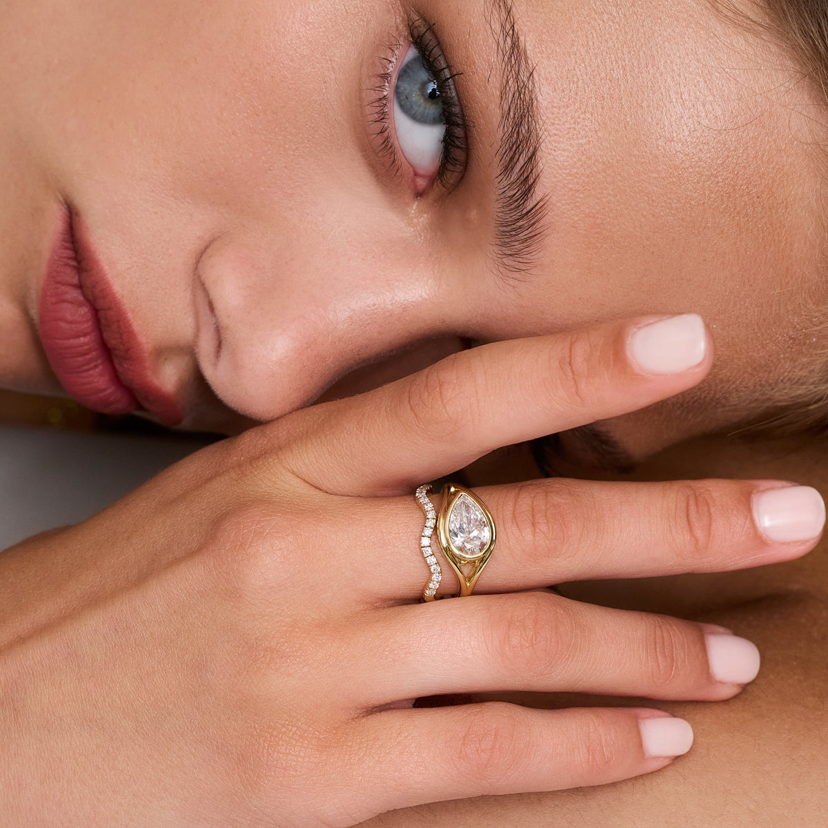 A close-up lifestyle shot of a woman's hand resting near her face, wearing a Pear shaped ring with a pear-cut diamond and a wave diamond ring.