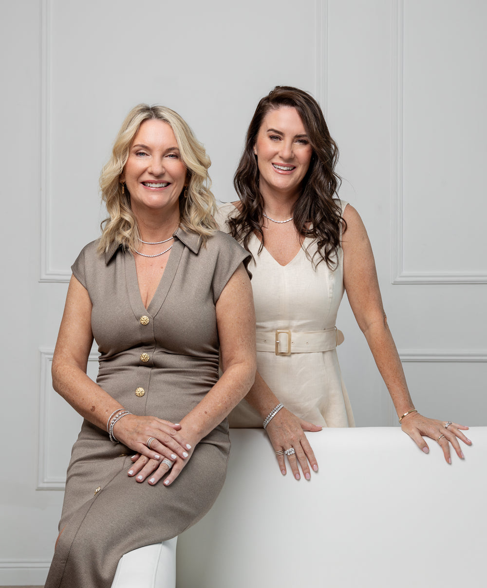 Two women wearing diamond jewellery sitting on a white chair against a light grey wall