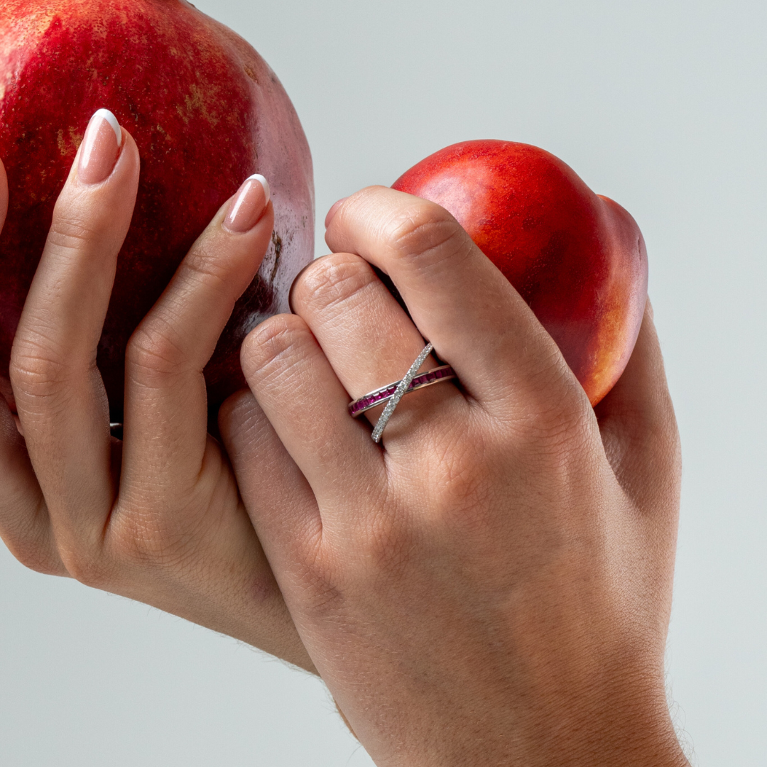Woman's hand holding apples showcasing Ruby Criss Cross ring with red rubies and diamonds gemstone ring.