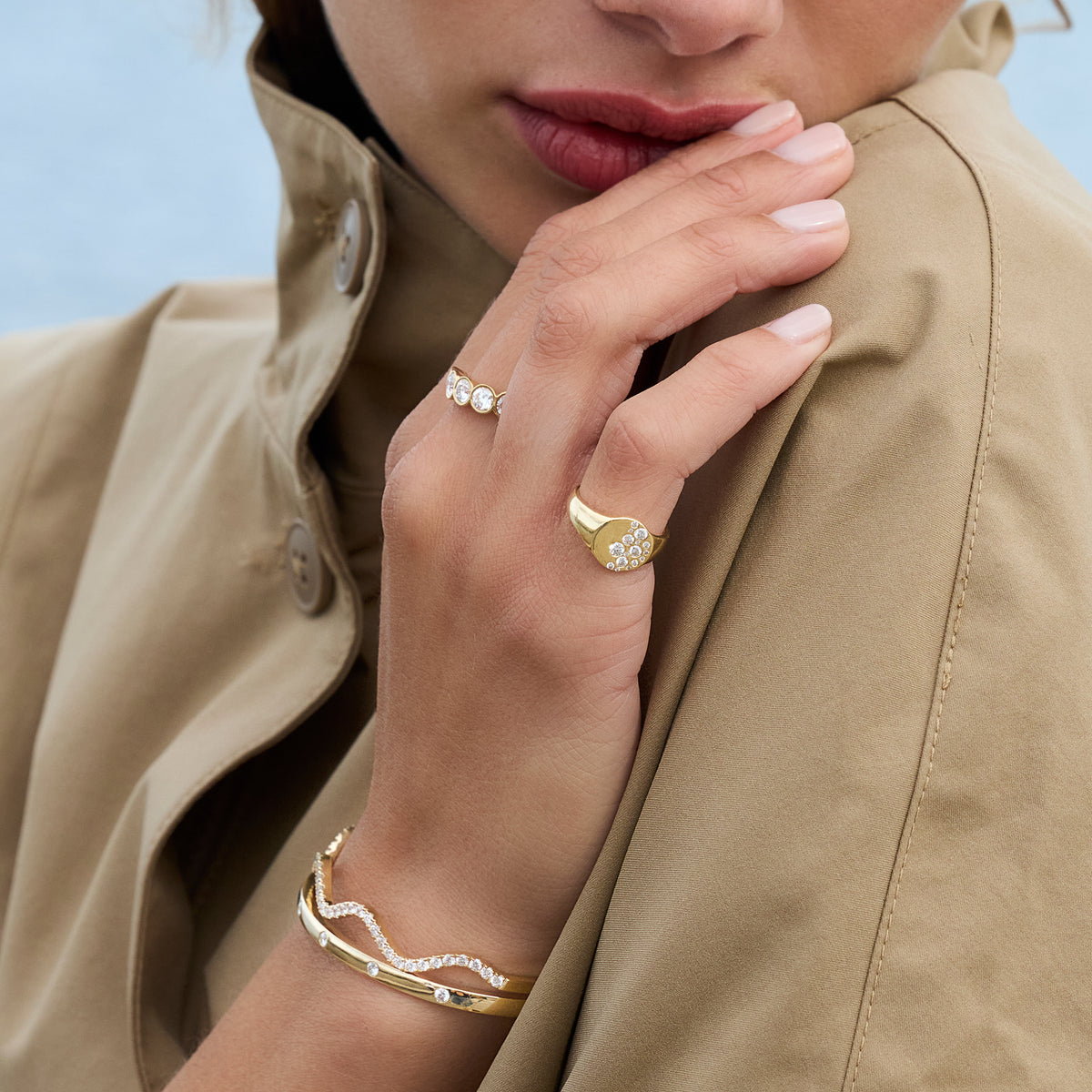 A close-up lifestyle shot of a model wearing gold jewelry, featuring a gold signet ring with round shape diamonds, a delicate gold wave bangle with diamonds, and another five stone bezel set diamond ring on her other hand, all styled with a beige trench coat against a soft blue background.