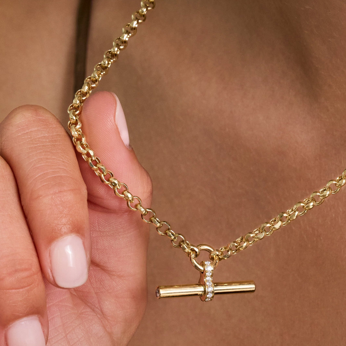 A close-up of a gold T bar necklace with small pave diamonds held by a hand against a warm skin-toned background.
