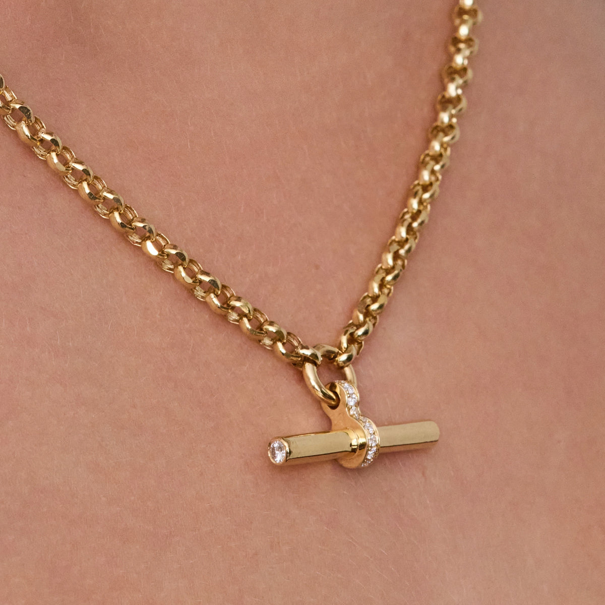 A gold T-bar necklace with a toggle clasp pendant, worn on a person's neck. The pendant features a horizontal bar with a small diamonds on each end and center.