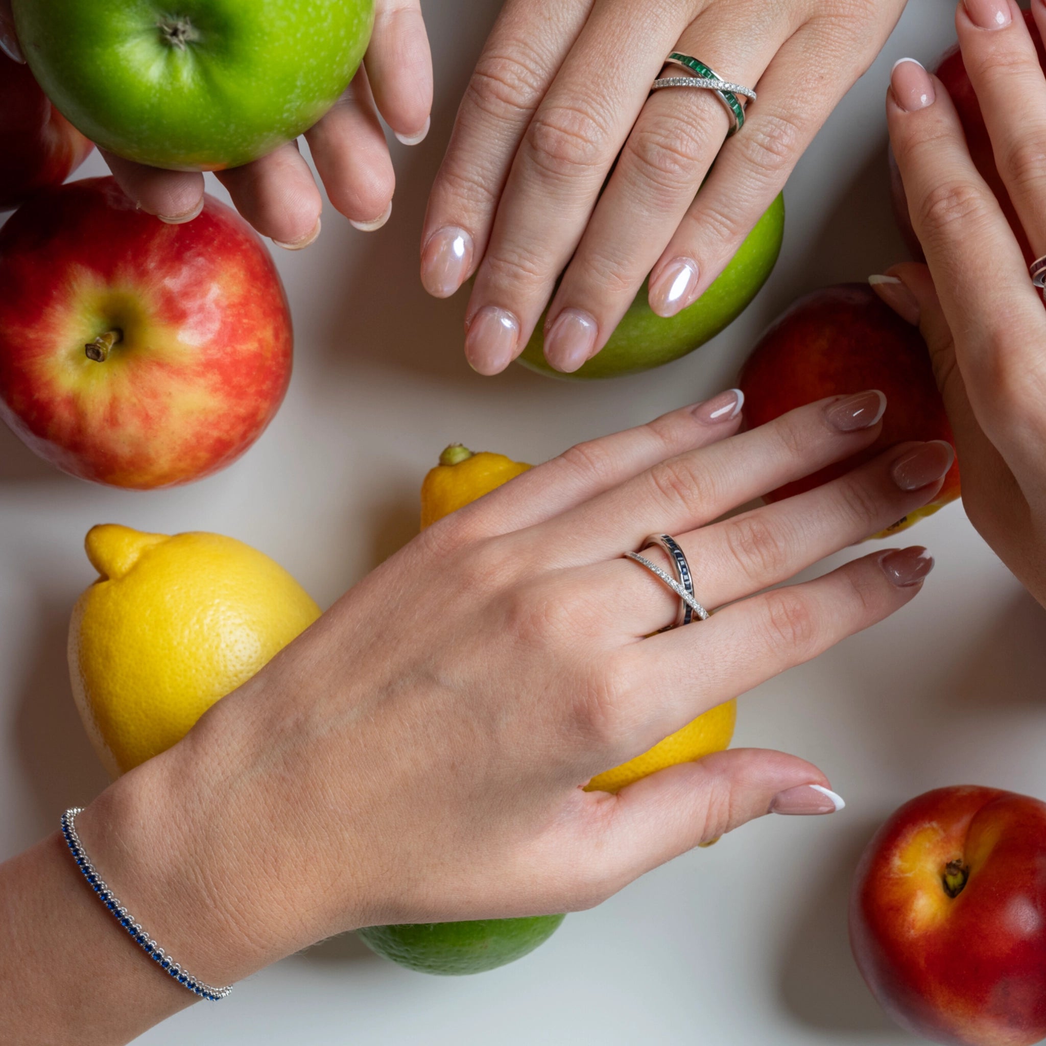 Two hands adorned with gemstone tennis bracelets and rings surrounded by colourful fruits on a white surface.