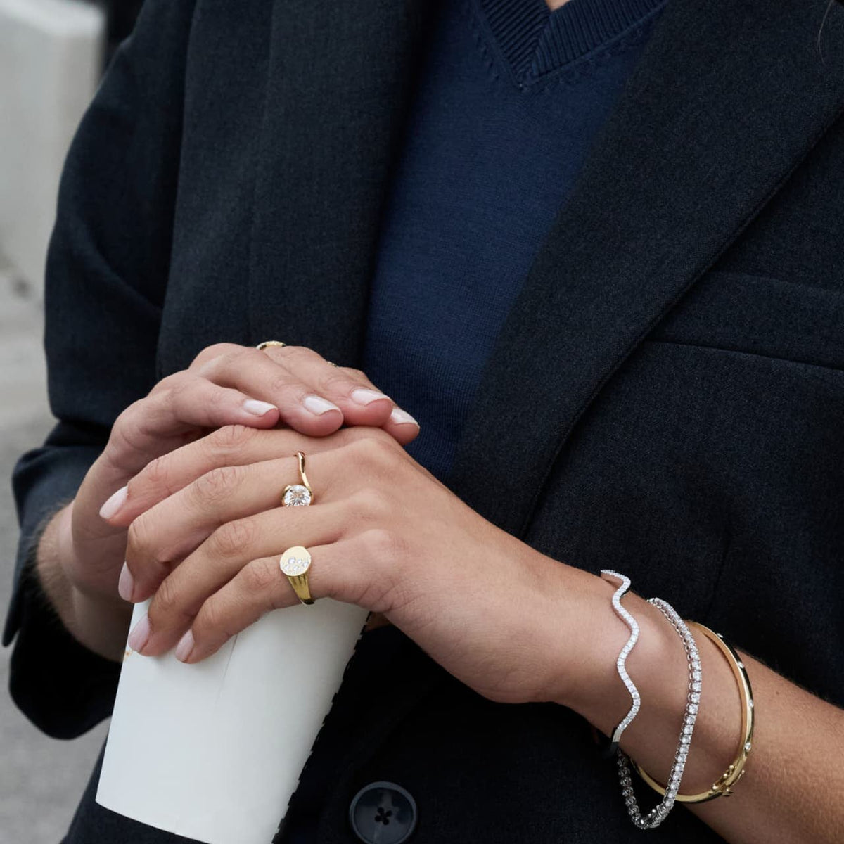 A close-up lifestyle shot of a person wearing a wavy diamond bracelet, a gold bangle, a gold signet ring, and a diamond ring, holding a white paper cup.