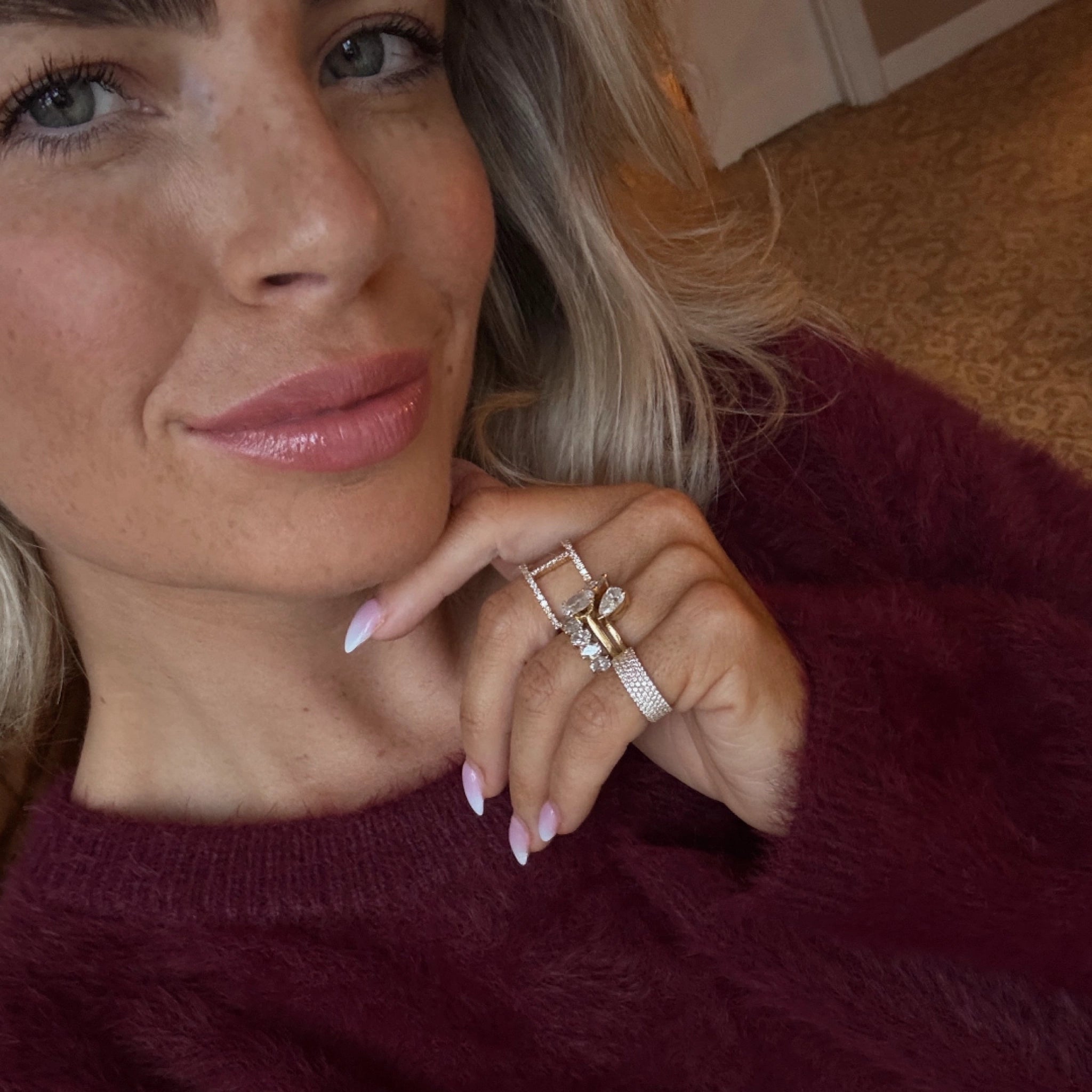 A woman showcases her stylish rings, including a four row pavé diamond ring, wearing a plush maroon sweater.
