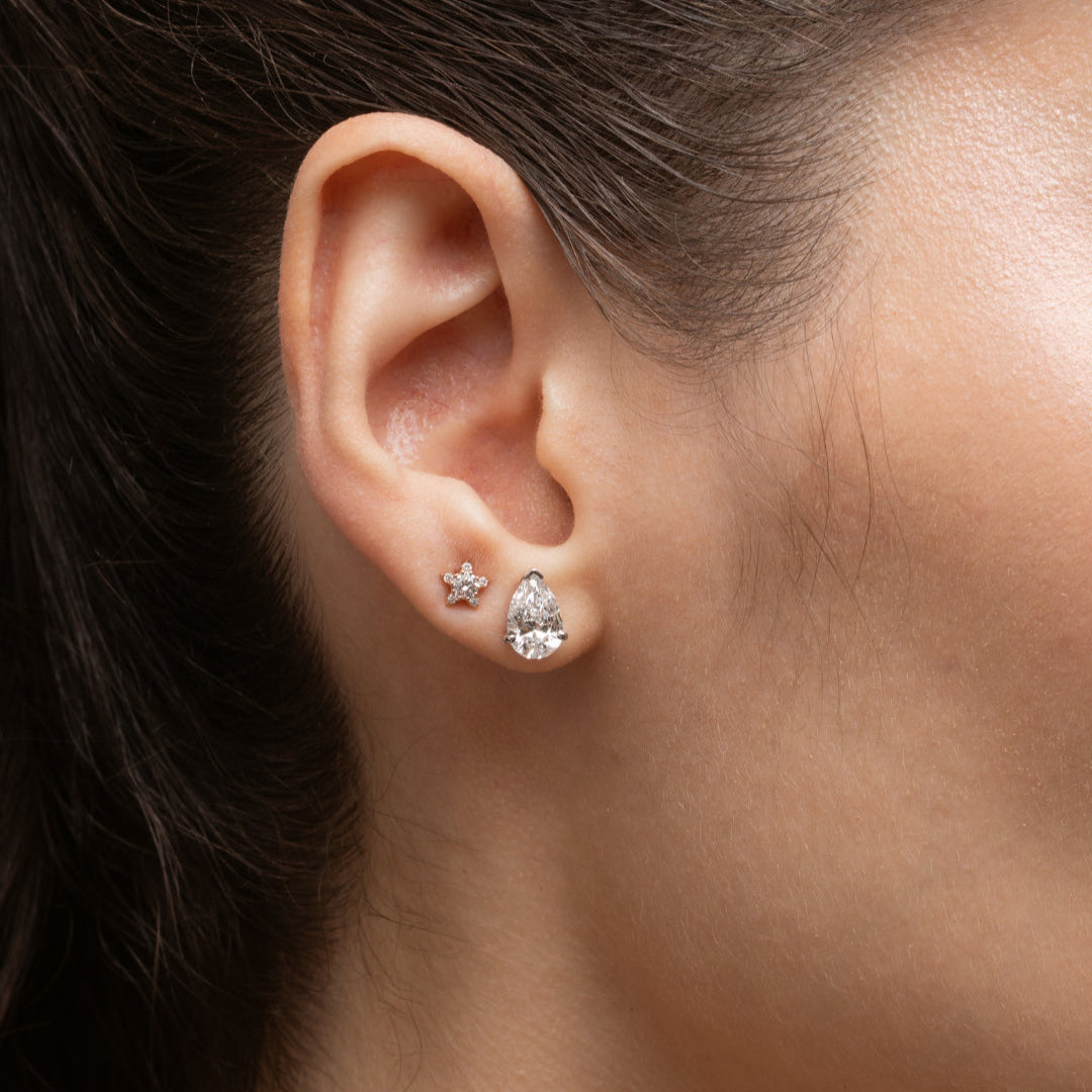 Pear Diamond Stud Earrings showcased in a woman's ear, featuring a sparkling pear shape diamond and a small star earring.