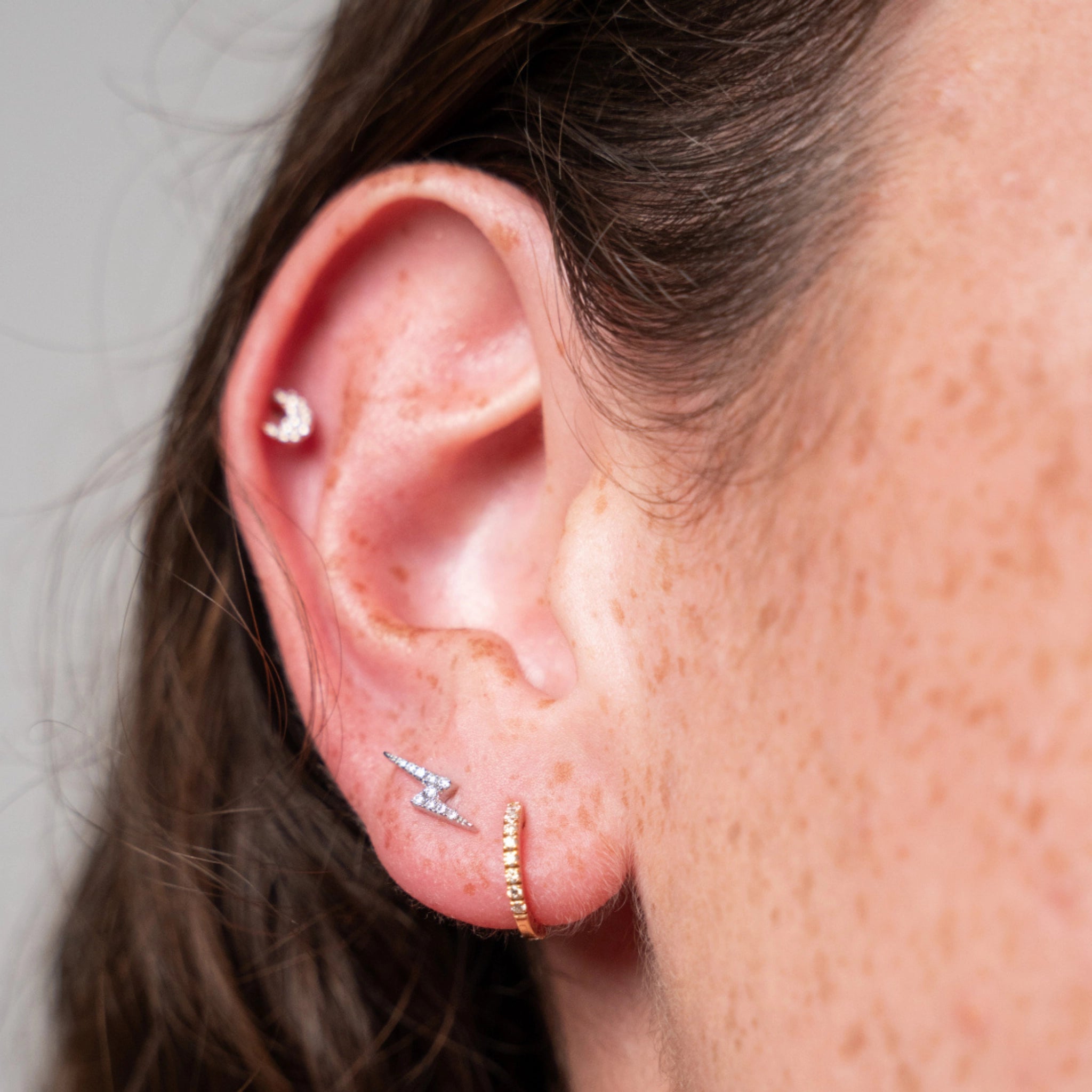 Close-up of a woman's ear with multiple earrings featuring the small moon earring against a neutral background