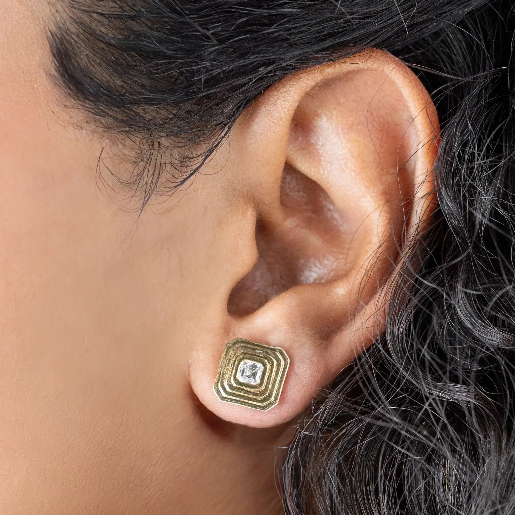 Close-up of a woman's ear wearing Tia earrings with asscher cut diamond, showcasing art deco style and gold design.